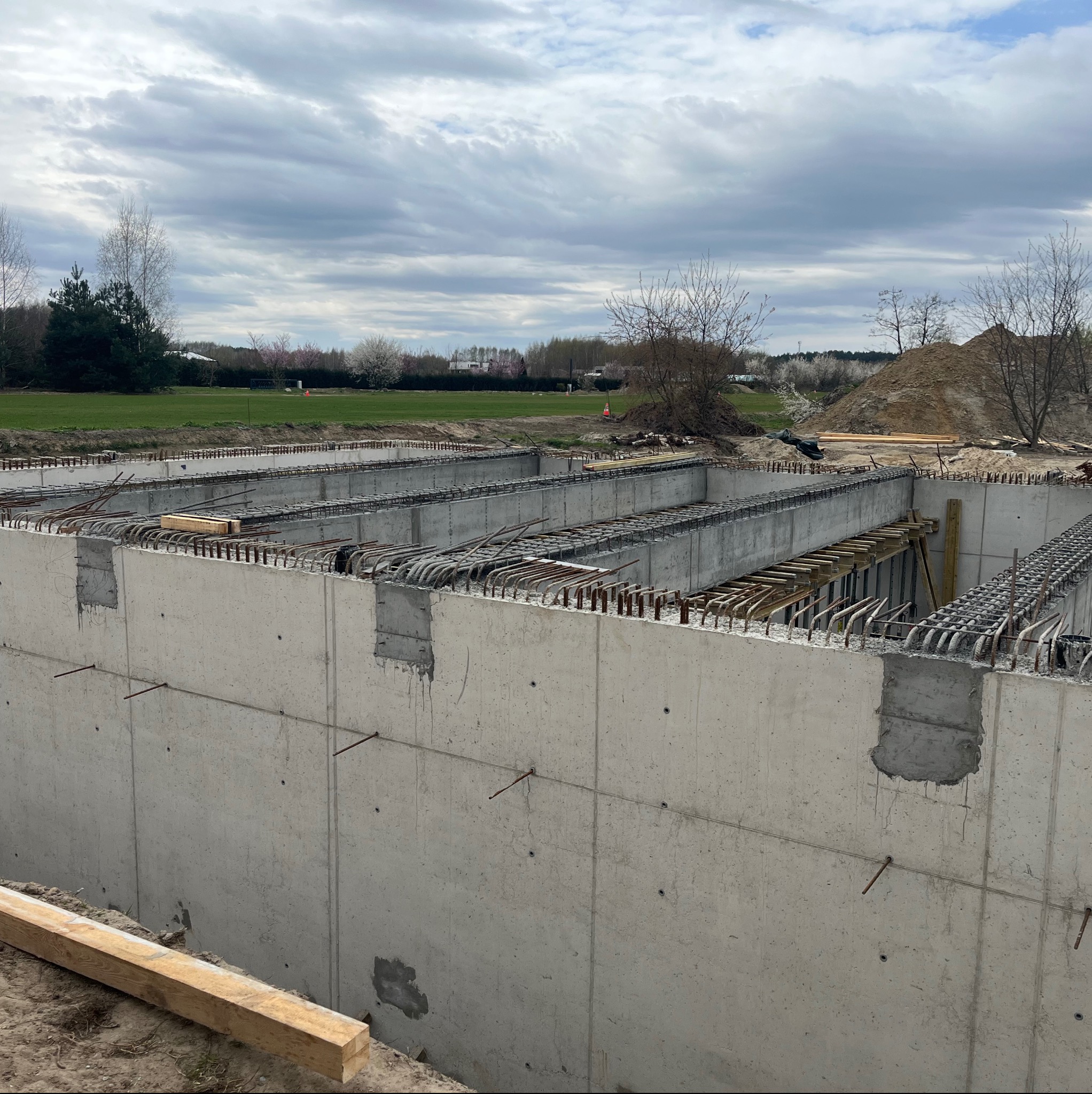 Żelbetowa konstrukcja fundamentów z widocznym zbrojeniem i szalunkiem, na tle zieleni i pochmurnego nieba. Etap budowy z bliska.