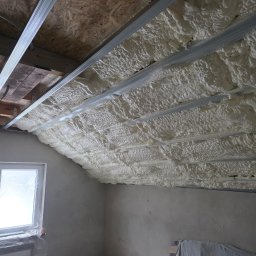 Poddasze z widoczną białą pianką izolacyjną między metalowymi profilami, okno z folią ochronną, surowe ściany. Detal z realizacji ocieplenia.