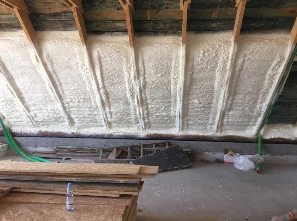 Poddasze po natrysku pianki PUR, widoczne drewniane krokwie. Na podłodze leżą płyty OSB i butelka wody. Izolacja wykonana z dbałością o detale.