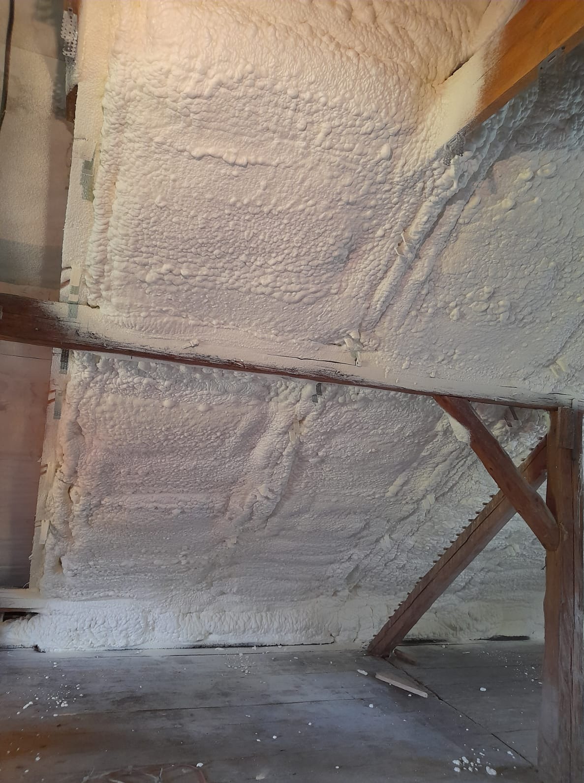 Poddasze z drewnianą konstrukcją pokryte grubą warstwą białej pianki izolacyjnej, widoczne nierówności i struktura piany, szara podłoga.