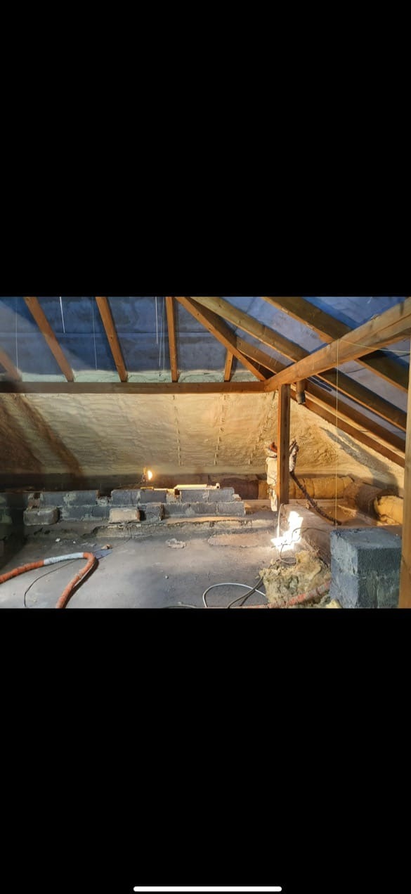 Poddasze z drewnianą konstrukcją dachu, częściowo pokryte pianką izolacyjną. Widoczne cegły i elementy instalacji. Oświetlenie punktowe podkreśla strukturę pianki.