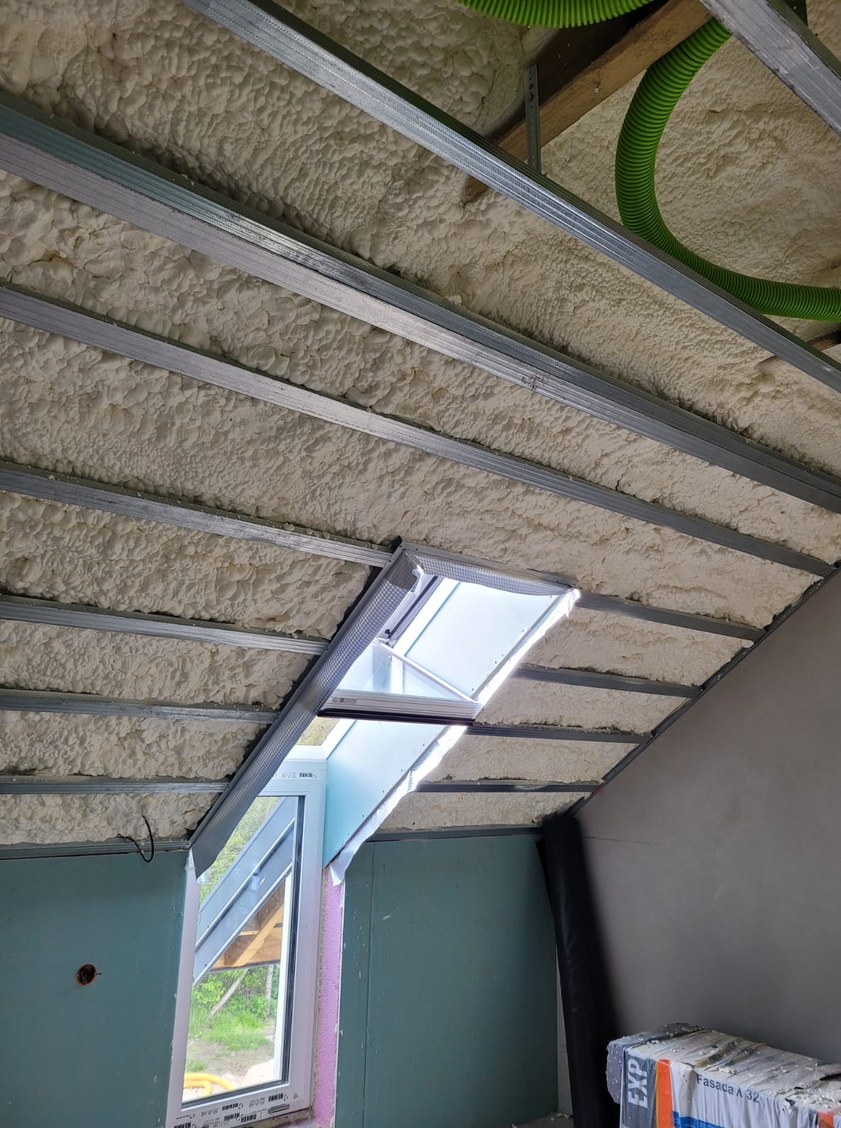 Poddasze z oknem dachowym po natrysku pianki PUR między metalowymi profilami. Widoczny fragment instalacji wentylacyjnej i ściany z płyt gipsowo-kartonowych.