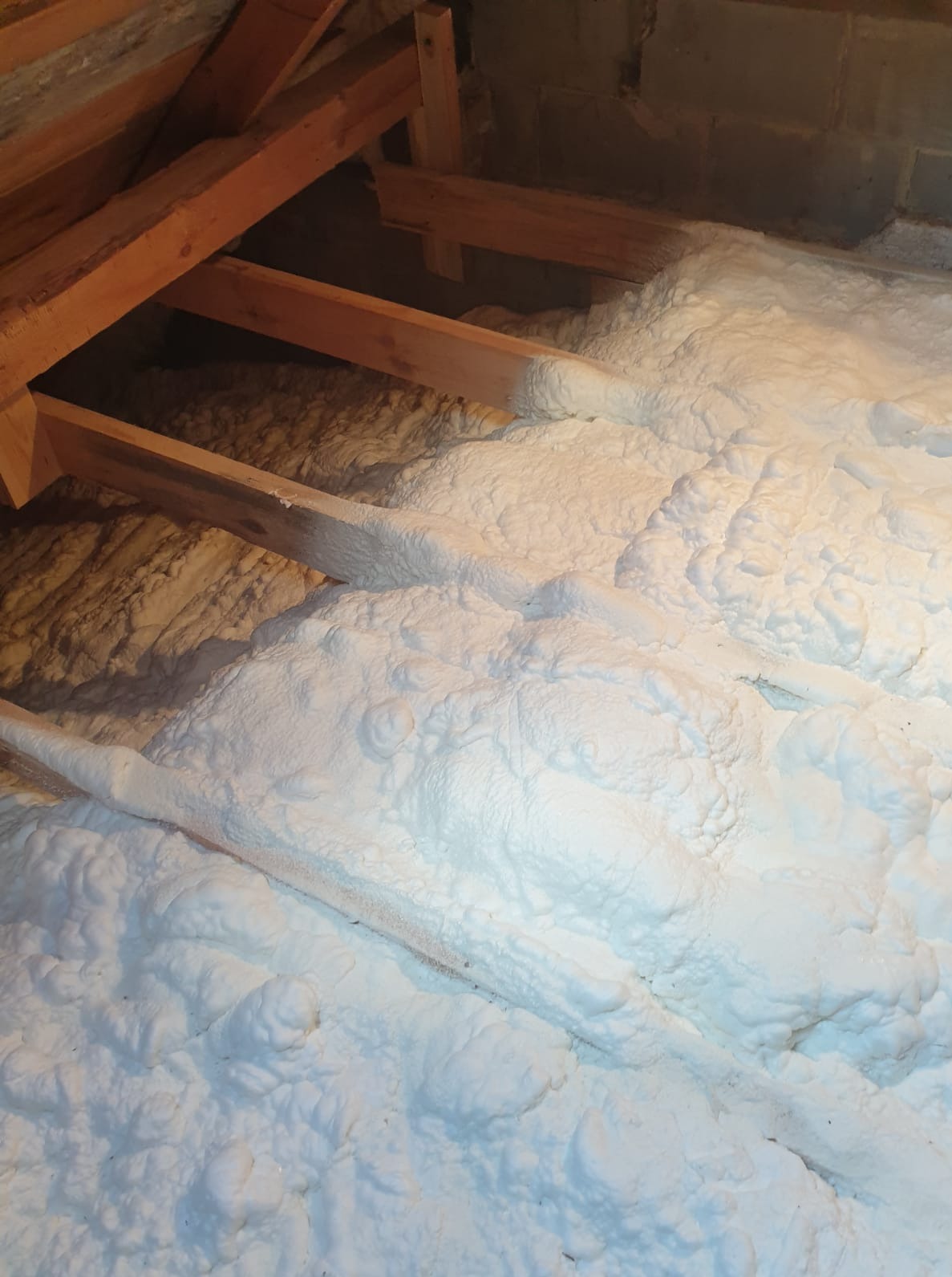 Poddasze z drewnianą konstrukcją i grubą warstwą białej pianki izolacyjnej, wypełniającą przestrzeń między krokwiami. Widoczne nierówności i struktura pianki.
