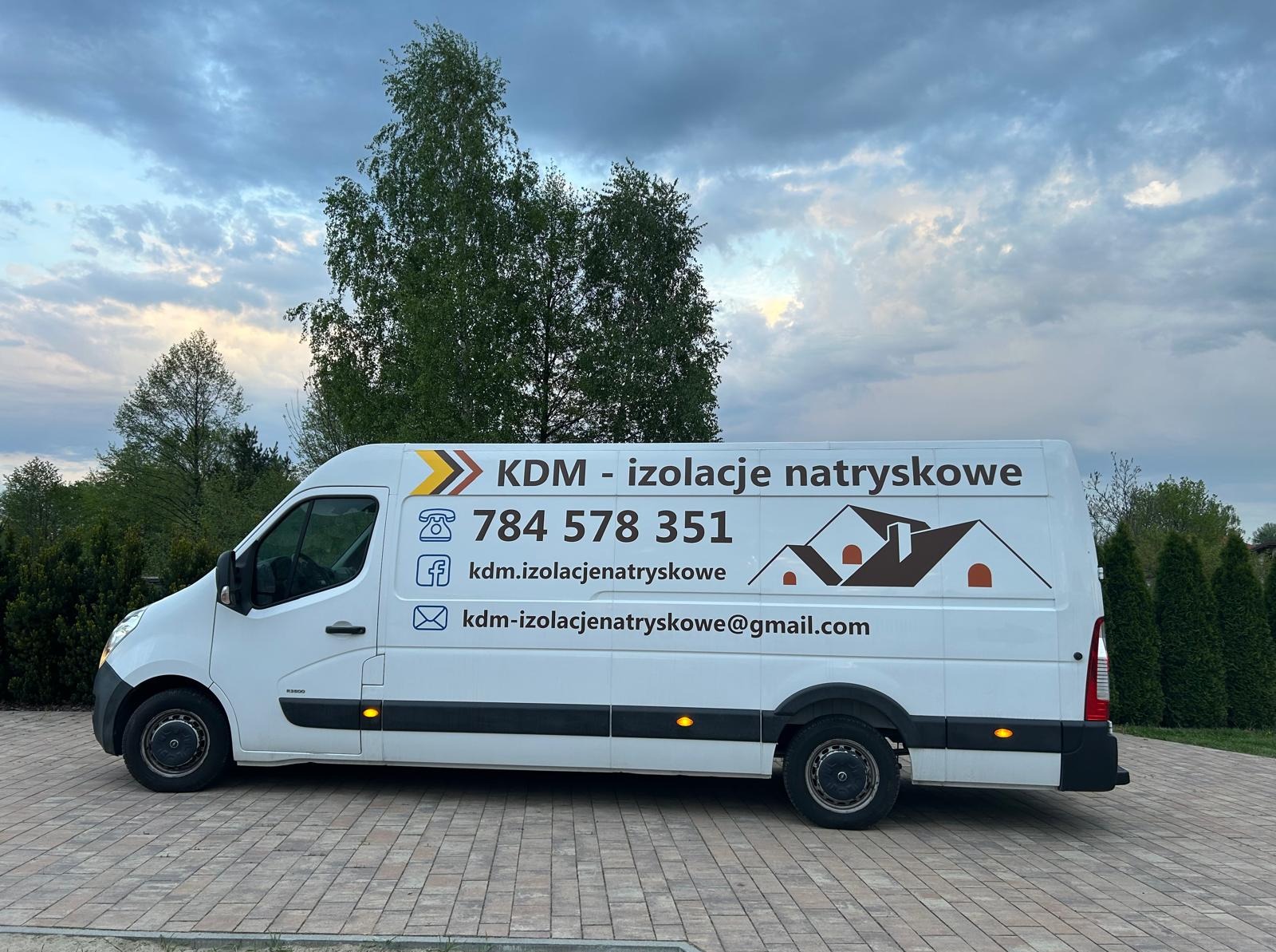 Biały van z logo firmy KDM - izolacje natryskowe, numerem telefonu i adresem e-mail na tle drzew i pochmurnego nieba. Reklama usług ocieplania.
