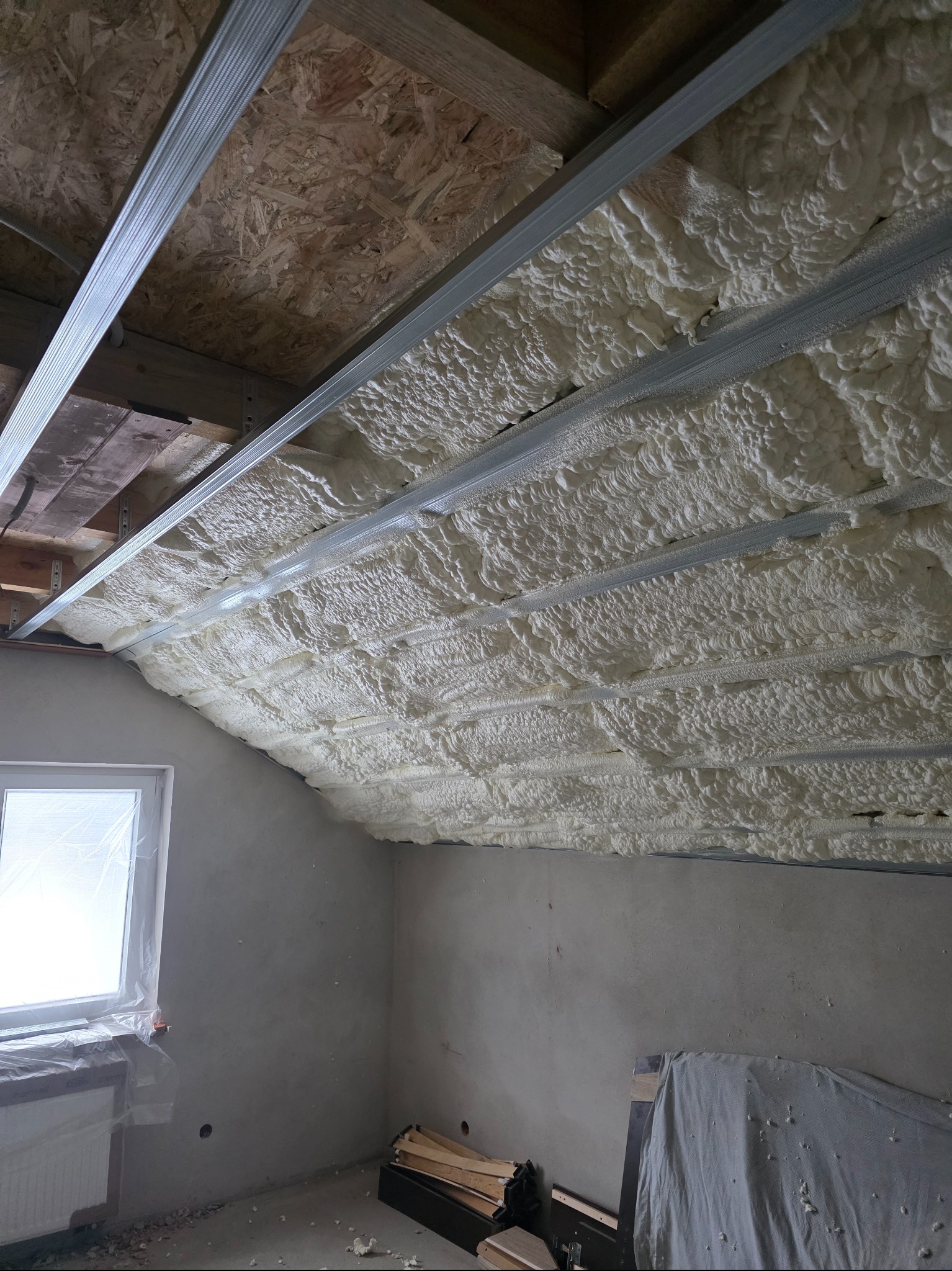 Poddasze z widoczną białą pianką izolacyjną między metalowymi profilami, okno z folią ochronną, surowe ściany. Detal z realizacji ocieplenia.