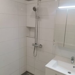 TOMASZ WĘGLARZ TITANIUM GROUP - Nowoczesna łazienka z białymi płytkami, deszczownicą i szarą podłogą. Minimalistyczny design z wnęką na kosmetyki i szafką pod umywalką.