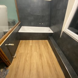 TOMASZ WĘGLARZ TITANIUM GROUP - Nowoczesna łazienka po remoncie: drewnopodobna podłoga, ciemne płytki na ścianach, biała wanna. Minimalistyczny design, widok z góry, ukończone wykończenie.