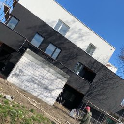 TOMASZ WĘGLARZ TITANIUM GROUP - Dwukondygnacyjny budynek z czarno-białą elewacją w trakcie prac wykończeniowych, widoczne rusztowania i pracownik w oddali, błękitne niebo w tle.