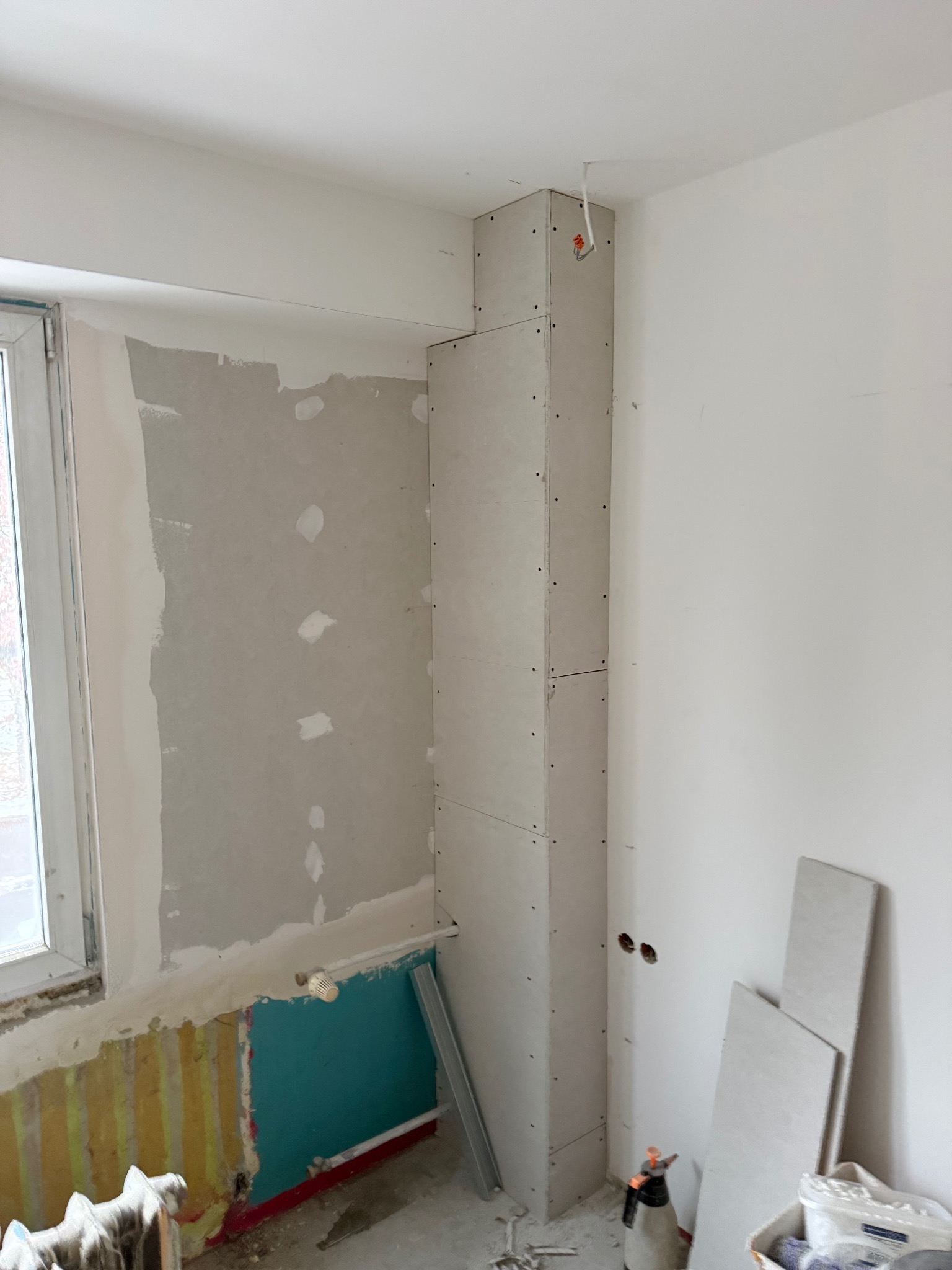 Wnętrze w trakcie prac: zabudowa z płyt karton-gips wokół pionu instalacyjnego przy oknie. Widoczne fragmenty starej ściany i przygotowania do dalszych prac wykończeniowych.