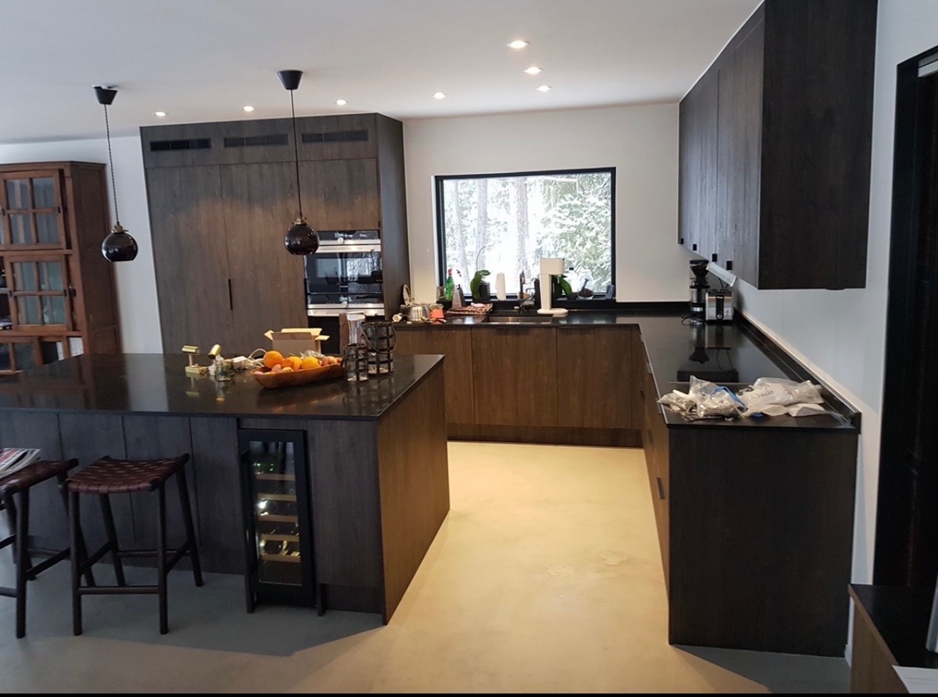 Nowoczesna kuchnia w Łętowem z ciemnymi drewnianymi szafkami, wyspą z winiarką i oknem z widokiem na zimowy krajobraz. Minimalistyczne lampy wiszące.