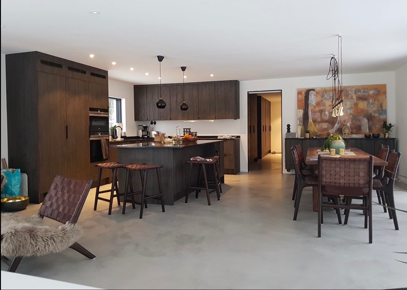 Nowoczesna kuchnia z wyspą i jadalnią w odcieniach brązu i szarości. Minimalistyczne lampy wiszące, drewniane krzesła i abstrakcyjny obraz dodają charakteru.