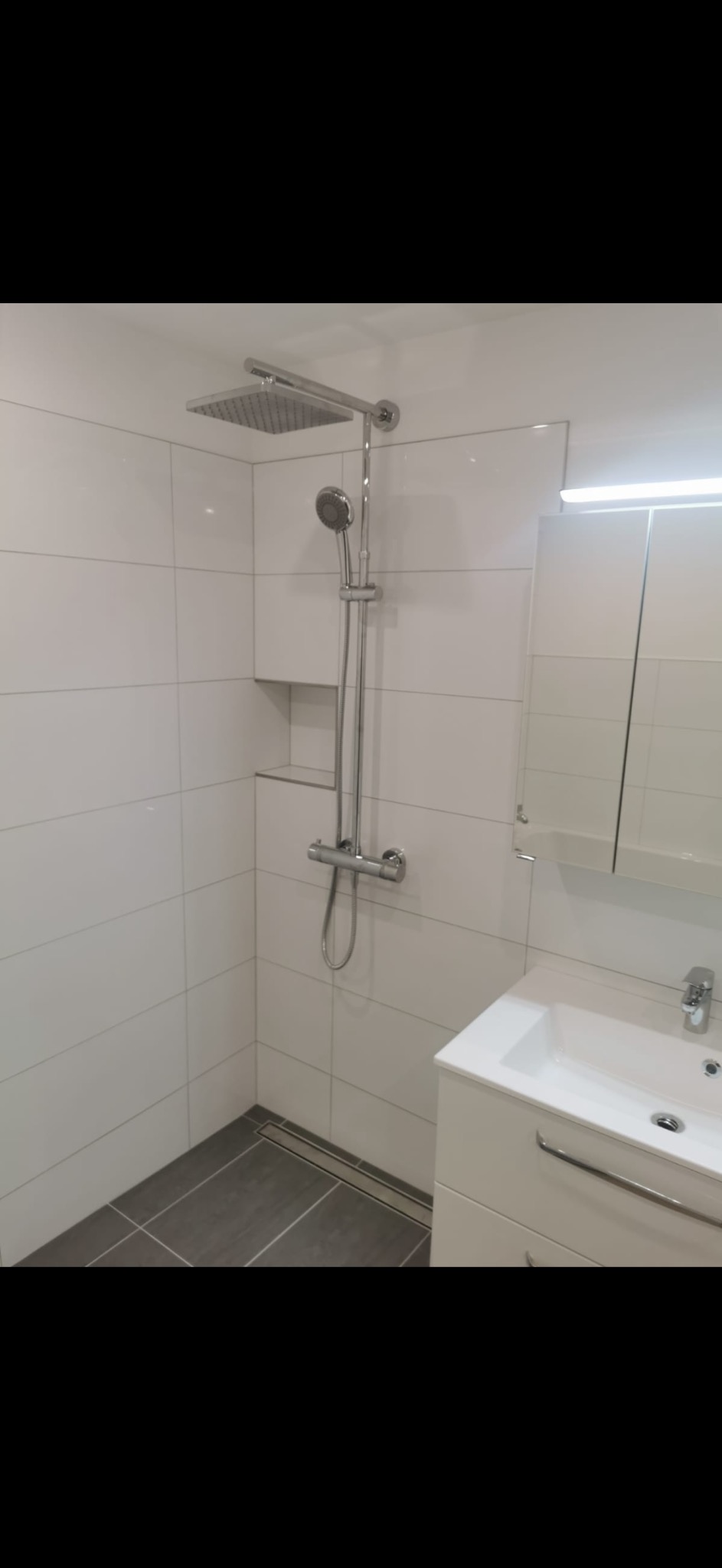 Nowoczesna łazienka z białymi płytkami, deszczownicą i szarą podłogą. Minimalistyczny design z wnęką na kosmetyki i szafką pod umywalką.