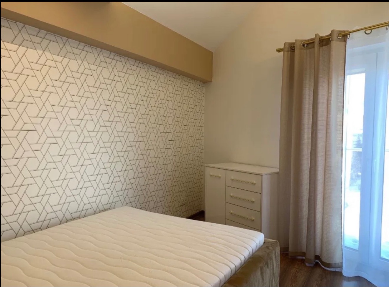 Pokój po remoncie: ściana z geometrycznym wzorem, beżowa listwa, biała komoda, zasłony w oknie. Widoczny fragment łóżka. Nowoczesny design.