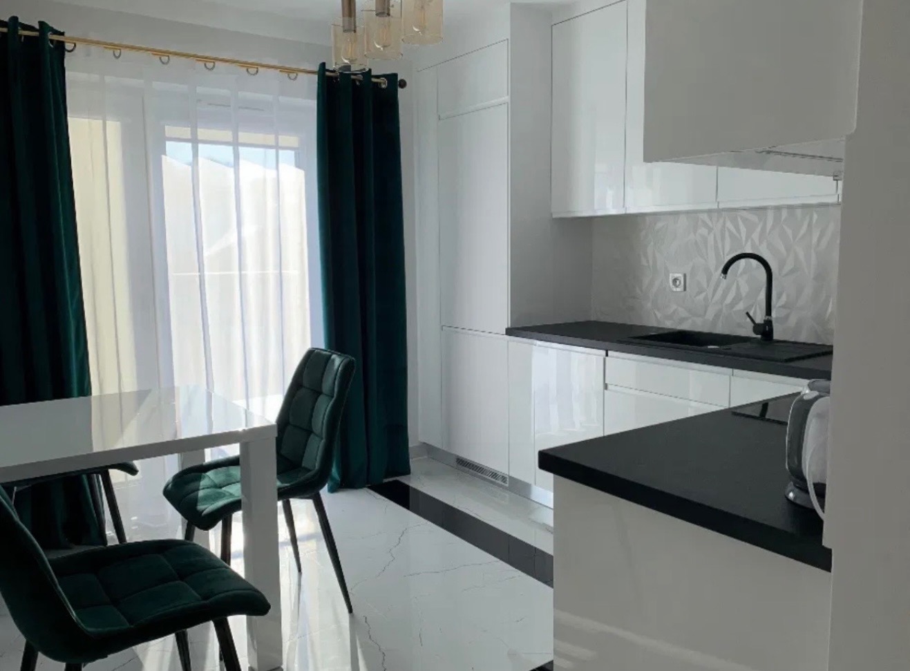 Nowoczesna, biała kuchnia z czarnymi akcentami: blat, zlew i pas podłogi. Szmaragdowe zasłony i krzesła dodają elegancji. Minimalistyczny design, jasne wnętrze.