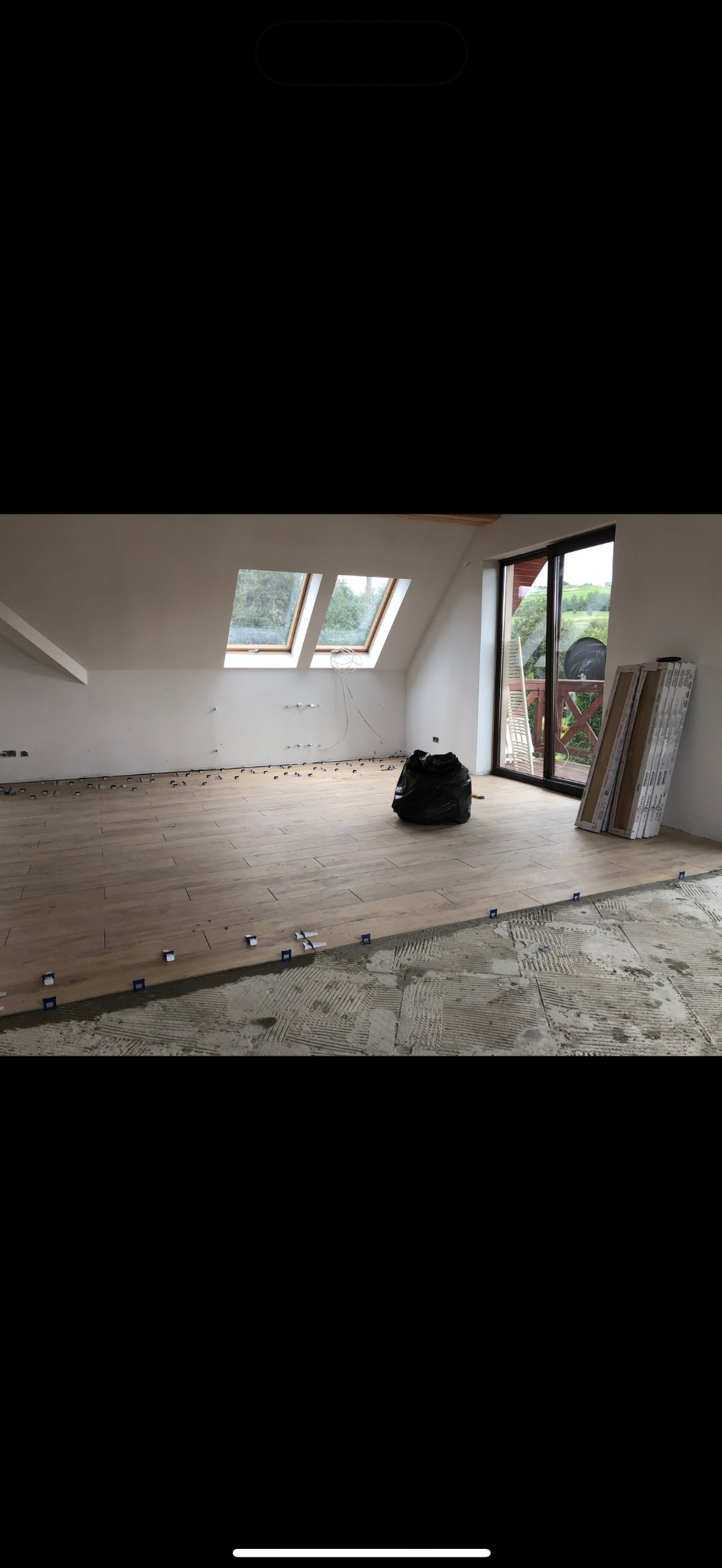 Podłoga z płytek imitujących drewno w trakcie układania na poddaszu. Widoczne krzyżyki dystansowe, częściowo położone płytki i przygotowane podłoże. Okno dachowe i drzwi balkonowe.