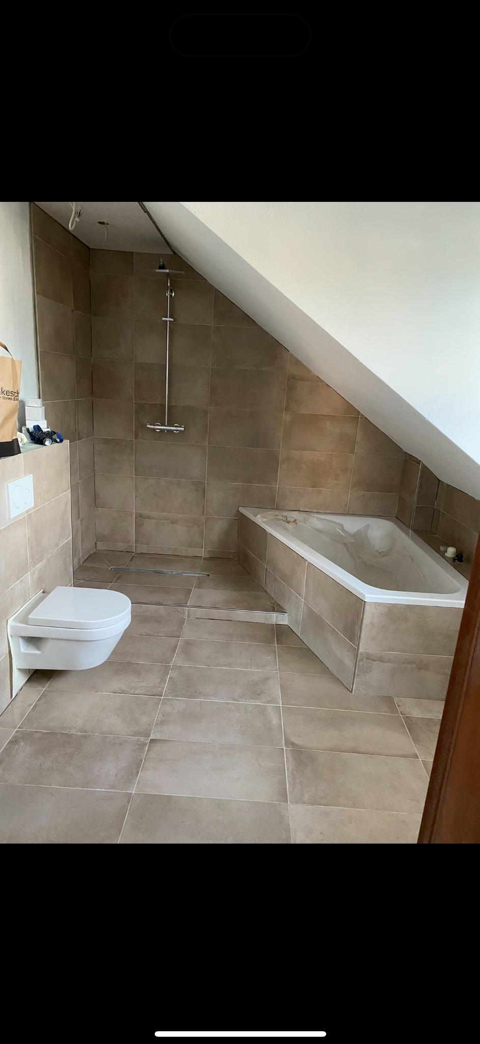 Wykończona łazienka na poddaszu z beżowymi płytkami, białą armaturą i kabiną prysznicową typu walk-in. Uwagę zwraca skośny sufit i minimalistyczny design.