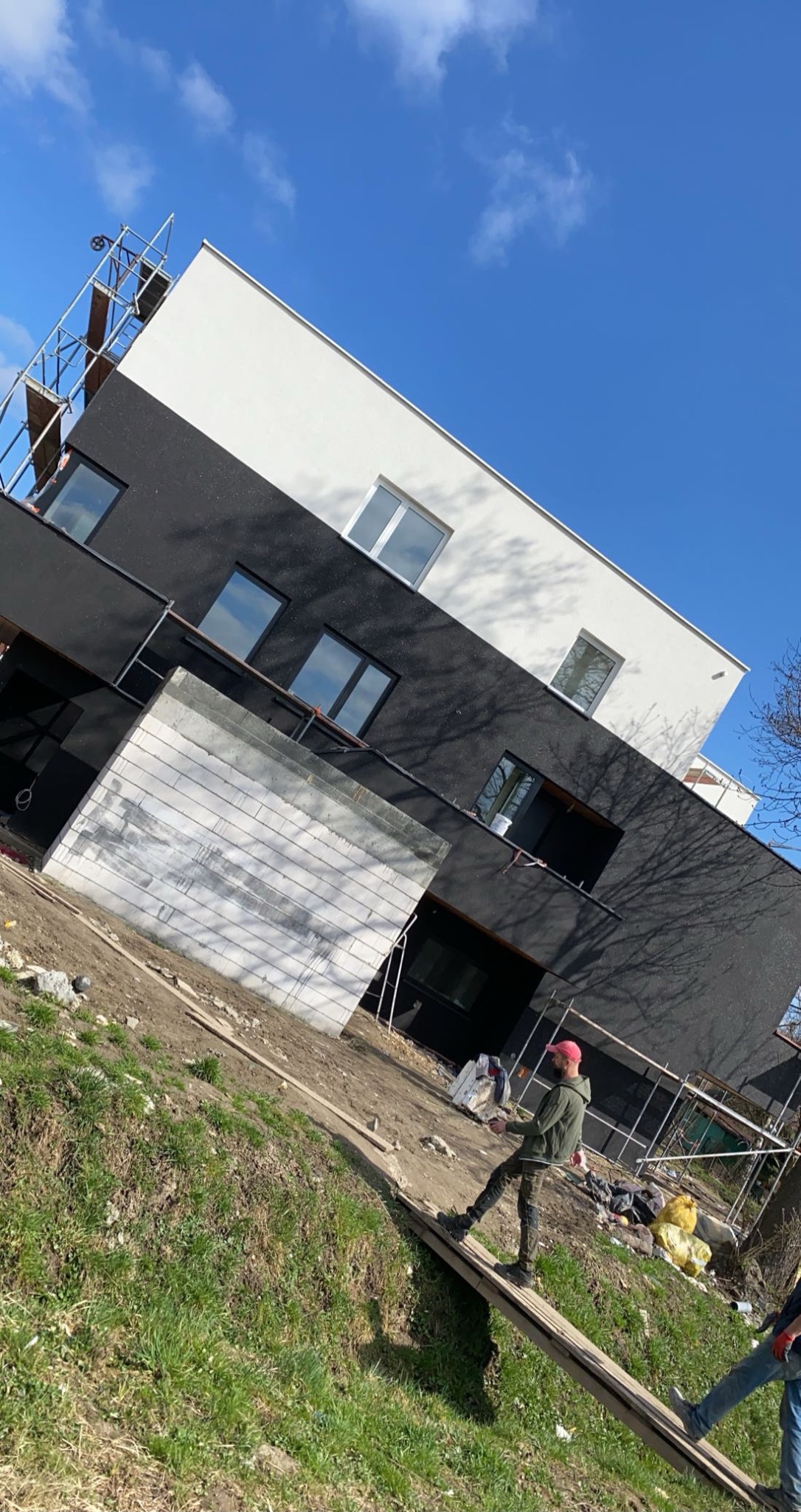 Dwukondygnacyjny budynek z czarno-białą elewacją w trakcie prac wykończeniowych, widoczne rusztowania i pracownik w oddali, błękitne niebo w tle.