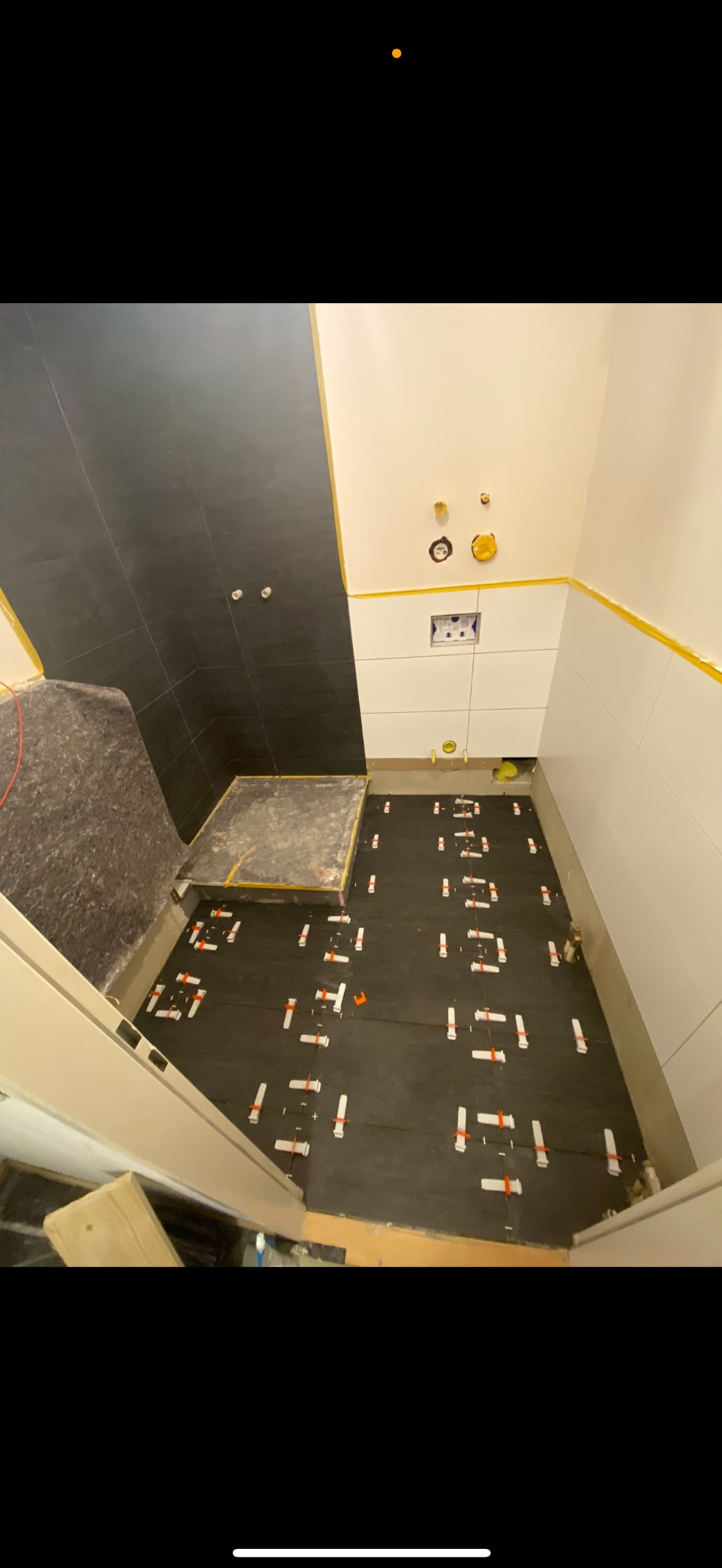 Łazienka w trakcie wykańczania: czarne i białe płytki na ścianach, podłoga z matą i systemem poziomowania płytek. Widoczne instalacje.