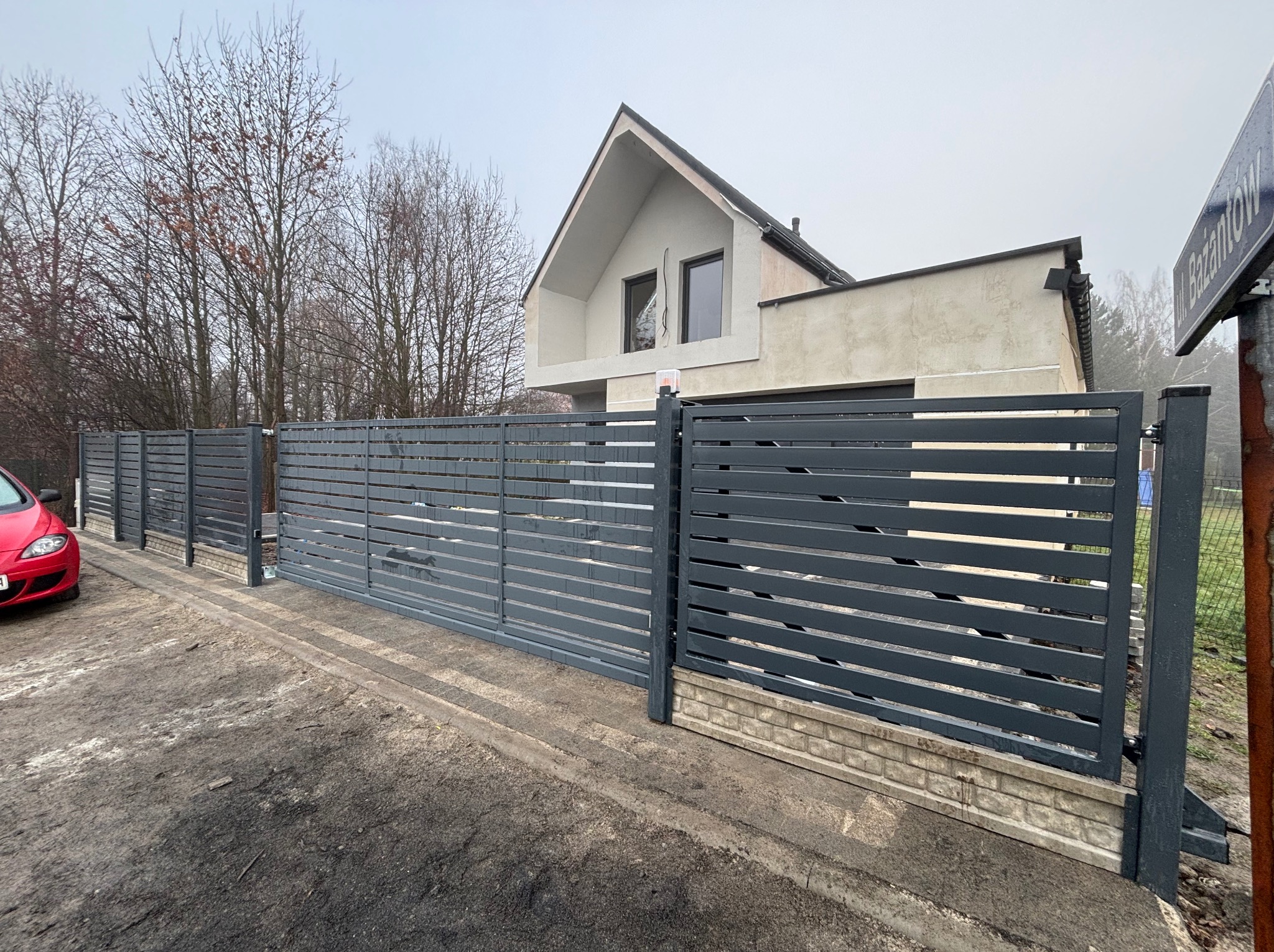 Nowoczesna, przesuwna brama wjazdowa z poziomych, szarych profili, na tle domu z dwuspadowym dachem i drzew bez liści. Widoczny fragment czerwonego auta.