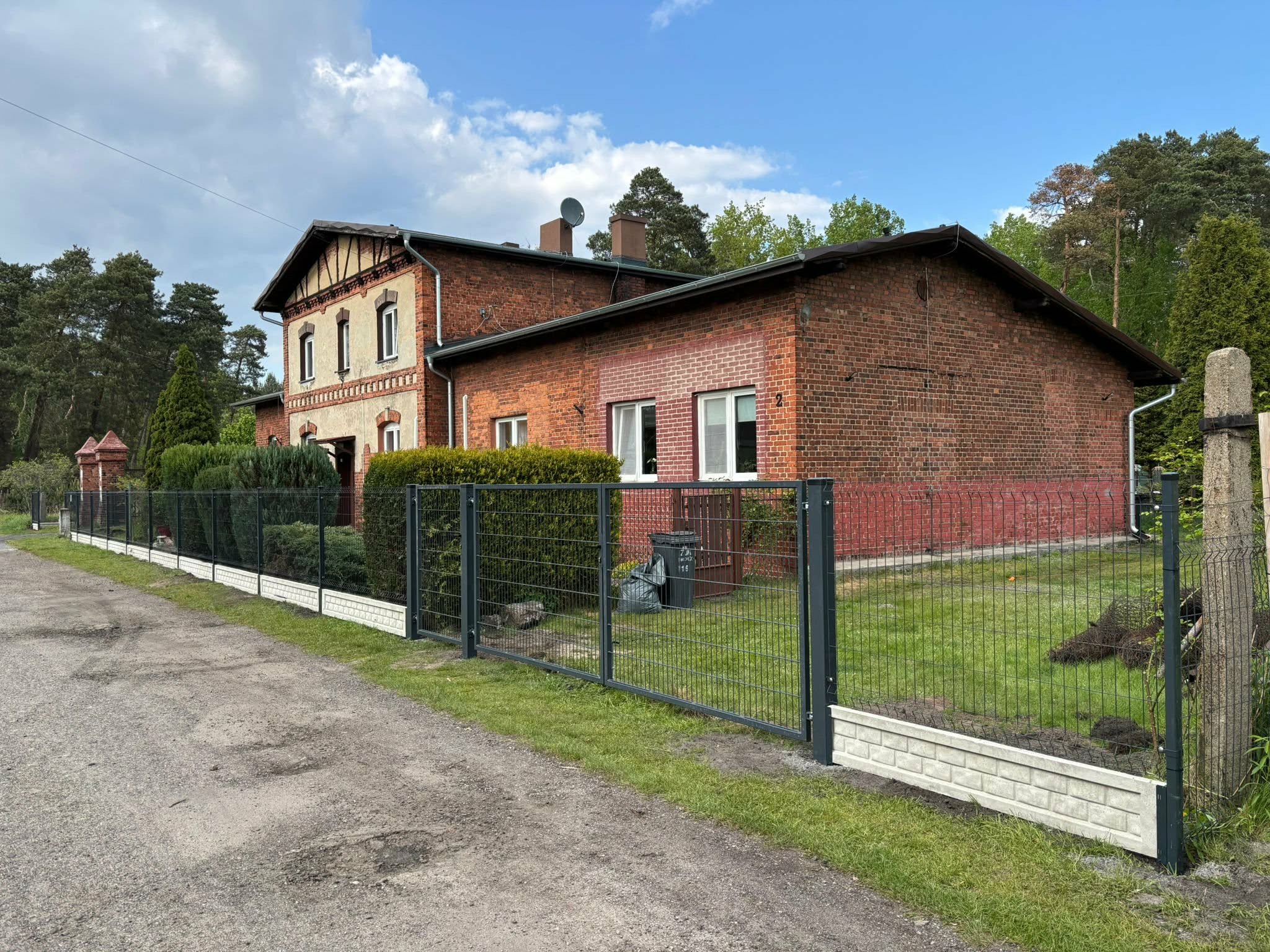 Nowoczesne ogrodzenie panelowe z podmurówką imitującą cegłę, otaczające posesję z ceglanym budynkiem. Widok z zewnątrz, w tle zieleń drzew i błękitne niebo.