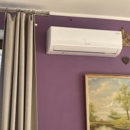 JAN Nizioł - Biała klimatyzacja na fioletowej ścianie w pokoju z zasłonami i obrazem w złotej ramie. Widok z dołu, oświetlenie dzienne.