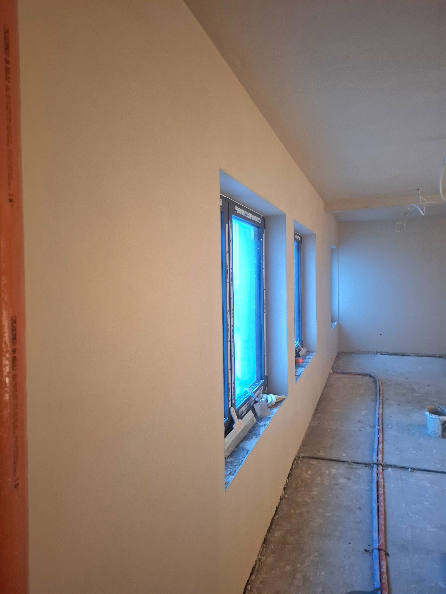 Wnętrze po tynkowaniu: gładkie, jasne ściany z trzema oknami zabezpieczonymi folią. Na podłodze widoczne rury instalacyjne i wiadro. Świeżo położone tynki.