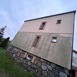 Dwukondygnacyjny budynek z elewacją w dwóch odcieniach beżu i kamienną podmurówką, widziany pod kątem z trawnika. Widoczne okna z brązowymi ramami i fragment dachu.