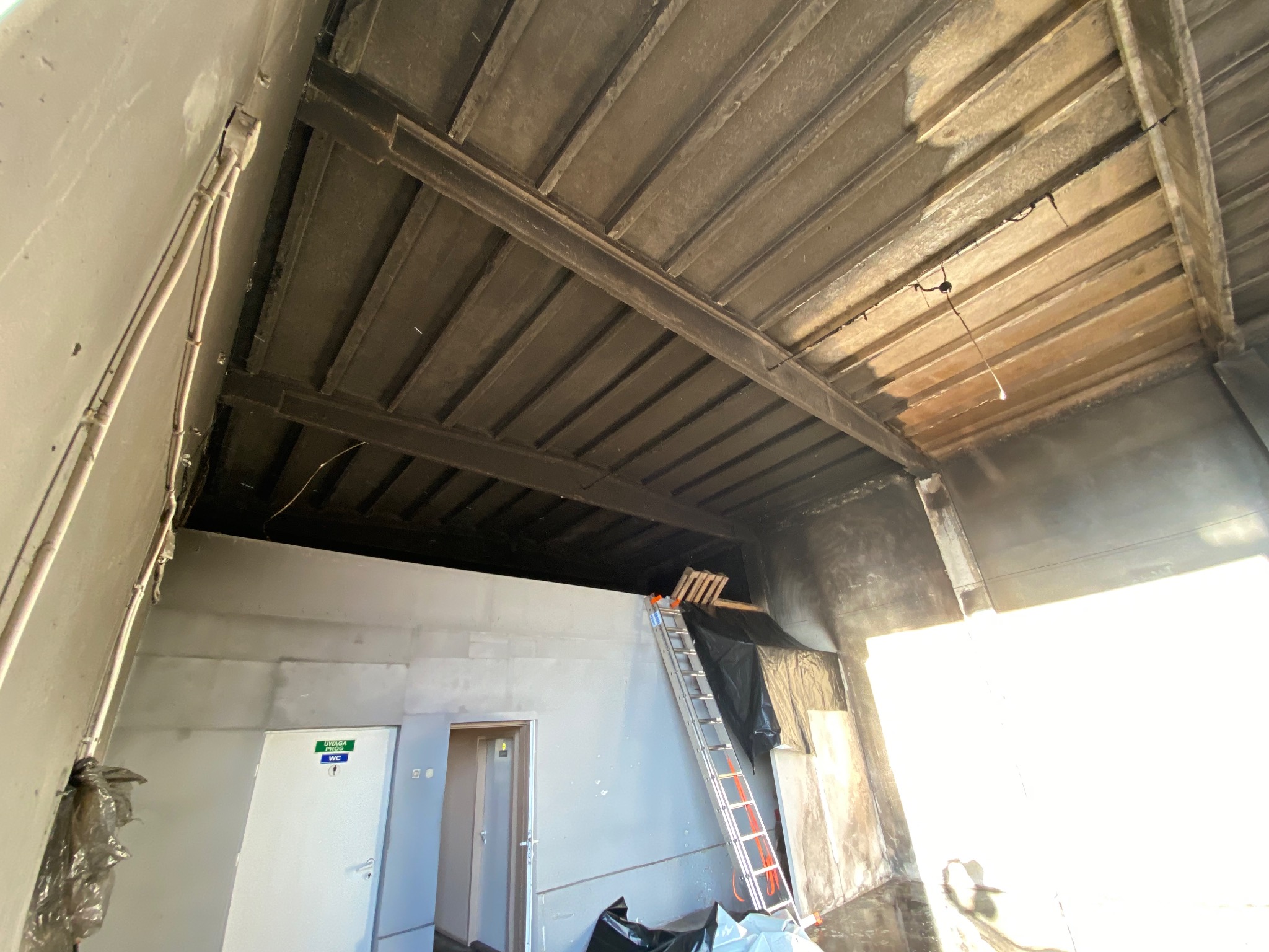 Wnętrze budynku w trakcie rozbiórki: widoczne elementy konstrukcyjne dachu, ściany, drabina oparta o ścianę, zabezpieczenia. Ujęcie z dołu, silne światło z prawej.