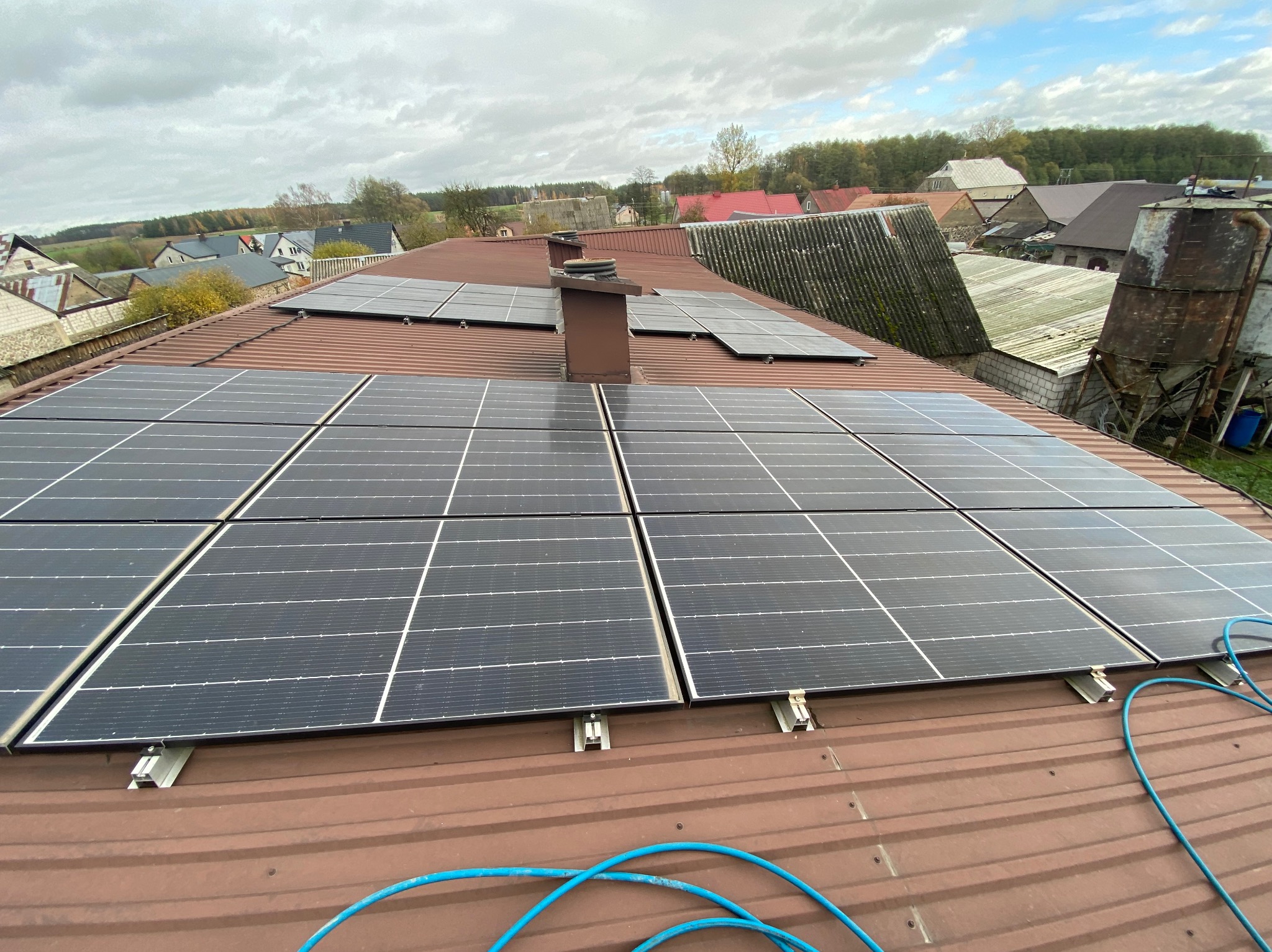 Panele fotowoltaiczne na brązowym, blaszanym dachu domu wiejskiego, widok z góry, pochmurne niebo. Częściowo widoczny niebieski przewód instalacyjny.