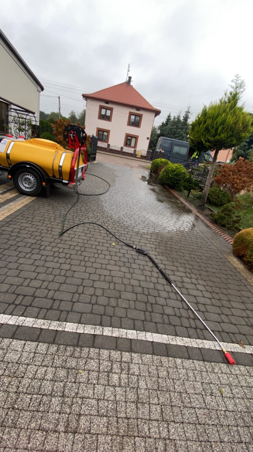 Żółty opryskiwacz na przyczepie i lanca na kostce brukowej, ślady wody. W tle dom jednorodzinny i krzewy. Usługa czyszczenia powierzchni zewnętrznych.