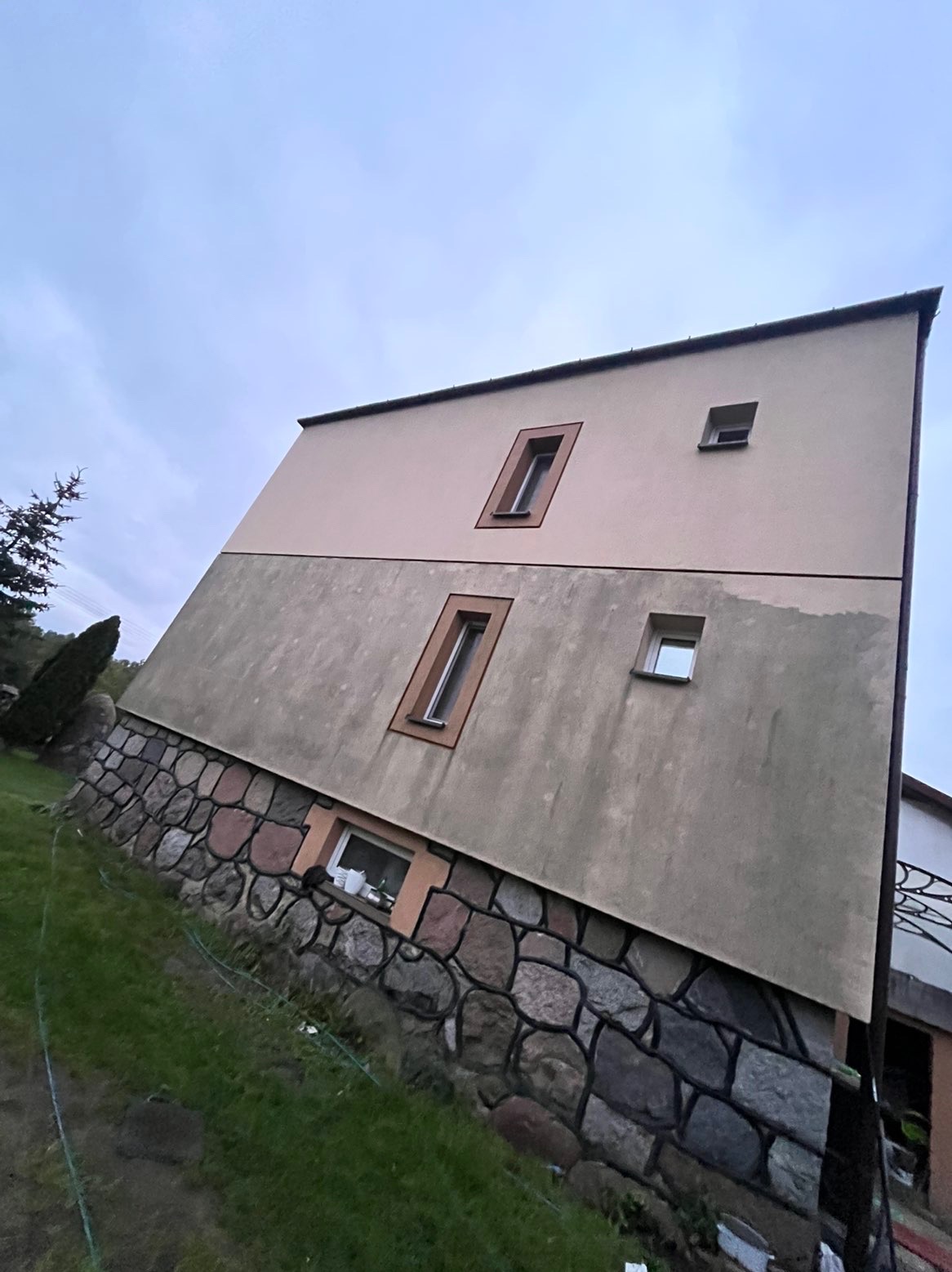 Dwukondygnacyjny budynek z elewacją w dwóch odcieniach beżu i kamienną podmurówką, widziany pod kątem z trawnika. Widoczne okna z brązowymi ramami i fragment dachu.