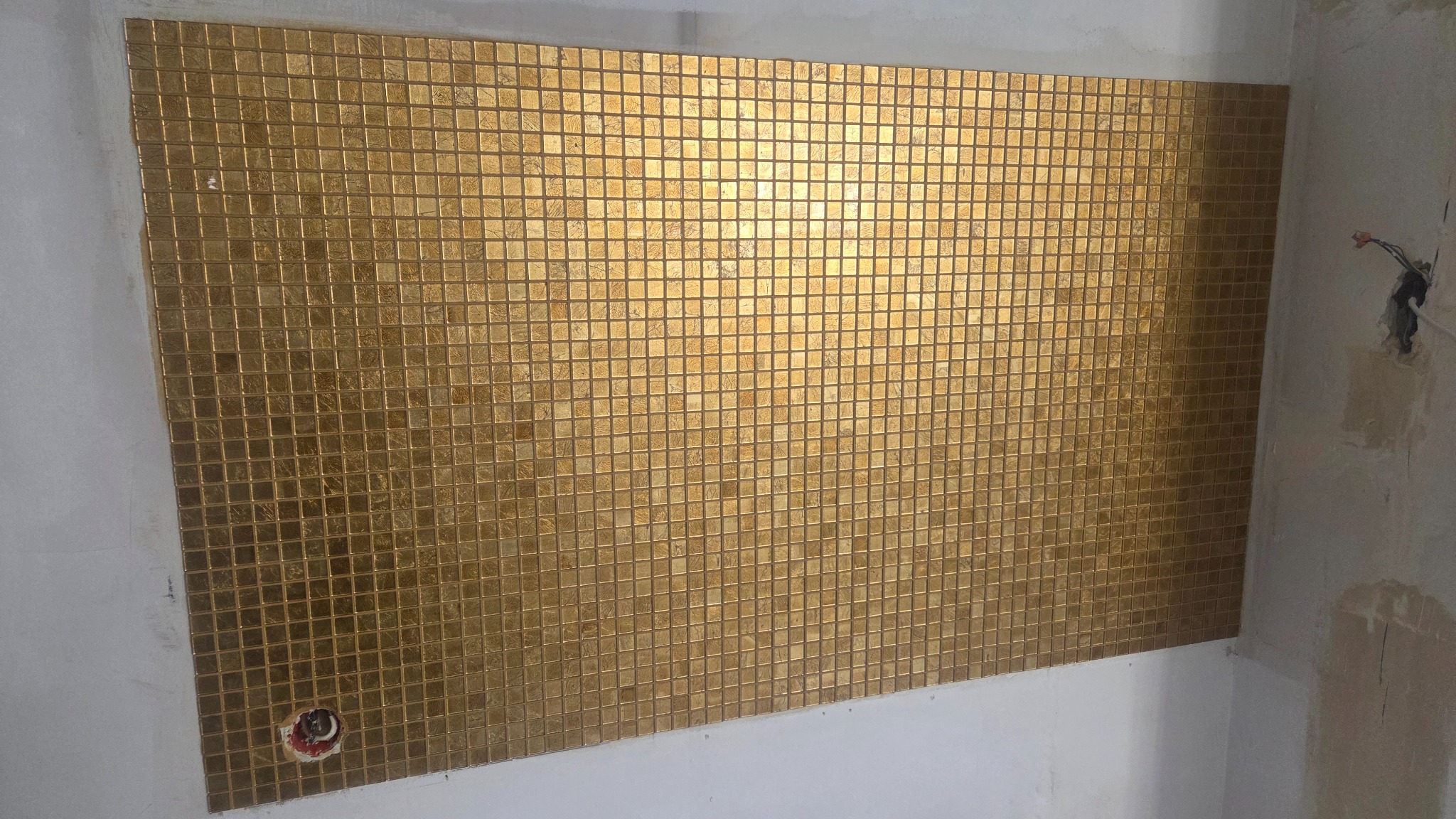 Ściana wyłożona złotą mozaiką w łazience, widoczne niedokończone prace wykończeniowe, w tle wystające kable elektryczne. Nietypowy, luksusowy wygląd.