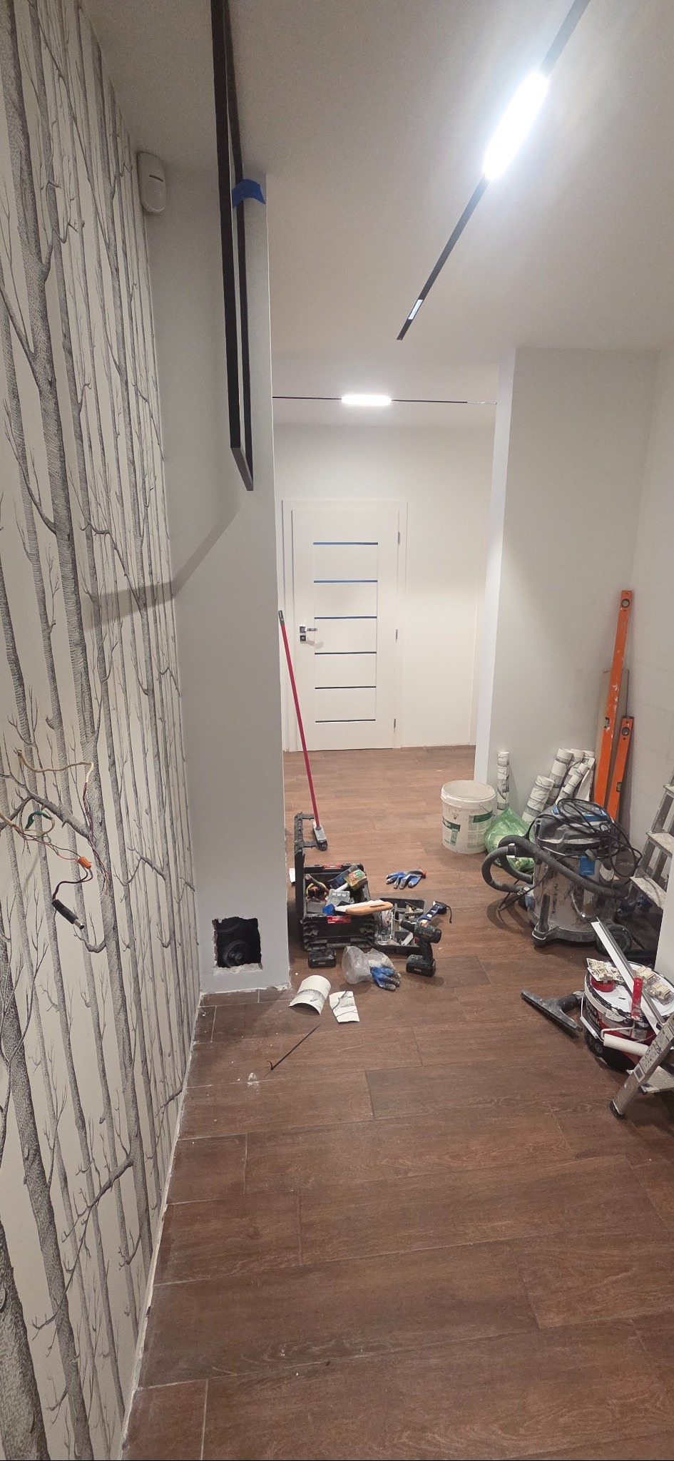 Korytarz w trakcie prac wykończeniowych: widoczne narzędzia, puszka elektryczna bez osprzętu, ściana z tapetą w motywie drzew i częściowo zamontowane oświetlenie.