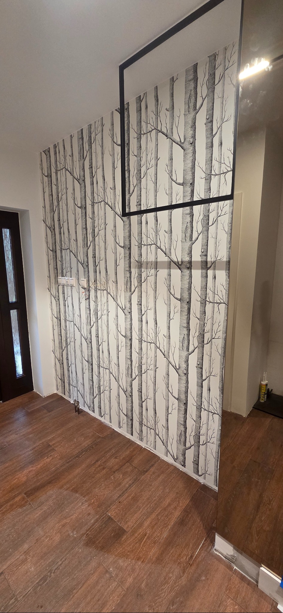 Wnętrze po remoncie: ściana z tapetą w motyw drzew, drewnopodobna podłoga, czarna rama lustra. Widoczny fragment drzwi wejściowych.