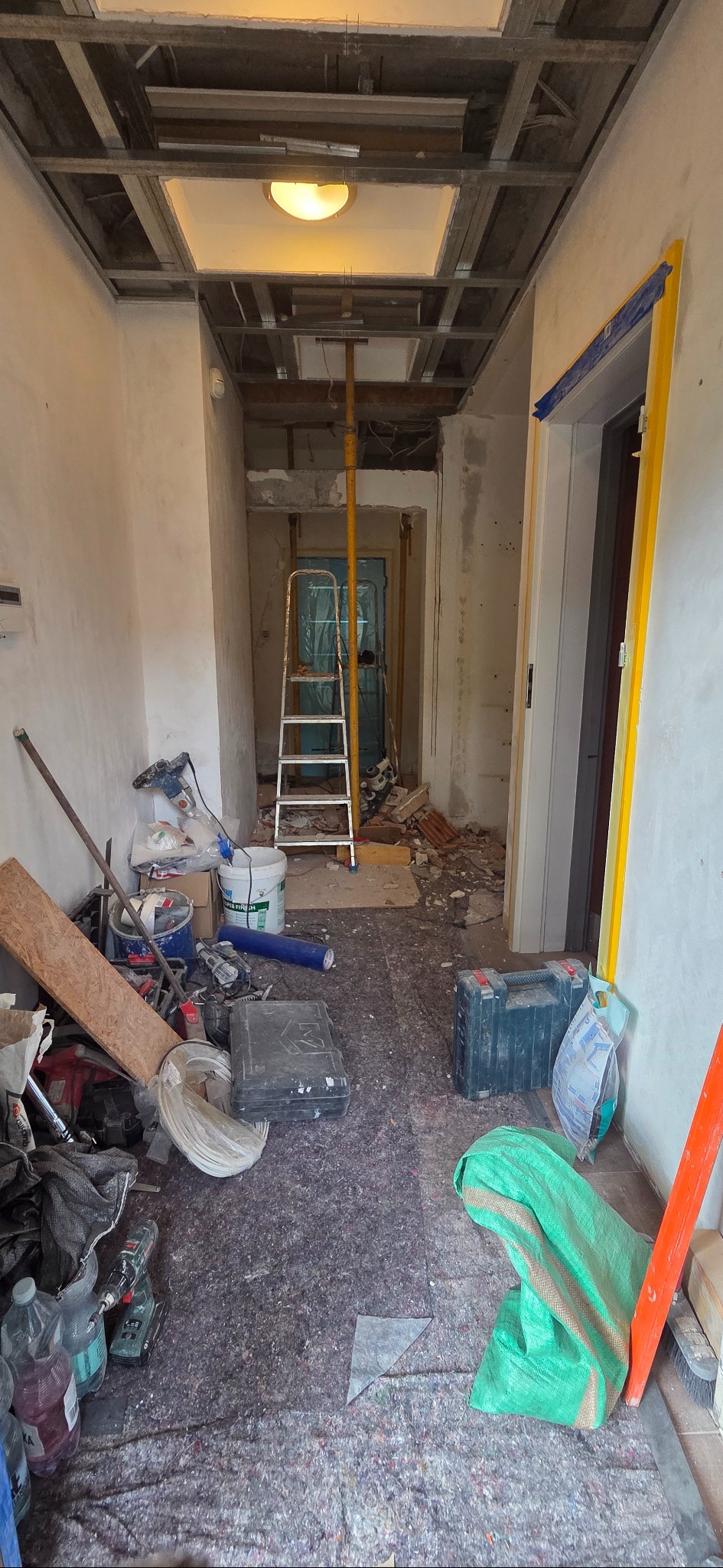 Wnętrze w trakcie remontu: widoczny sufit podwieszany w trakcie montażu, drabina, narzędzia i materiały budowlane. Otwór drzwiowy zabezpieczony folią.