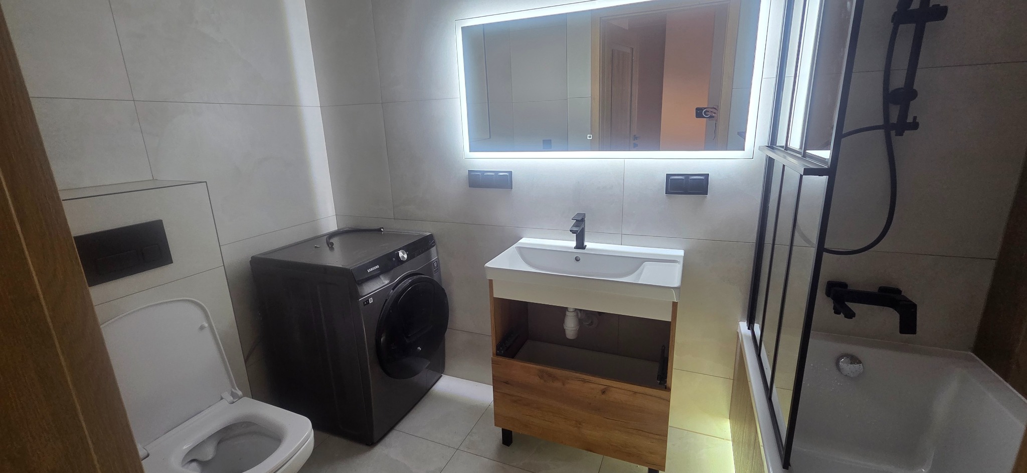 Nowoczesna łazienka z pralką, umywalką z szafką w odcieniach drewna i podświetlanym lustrem. Minimalistyczny design, szare płytki, czarna armatura i kabina prysznicowa.