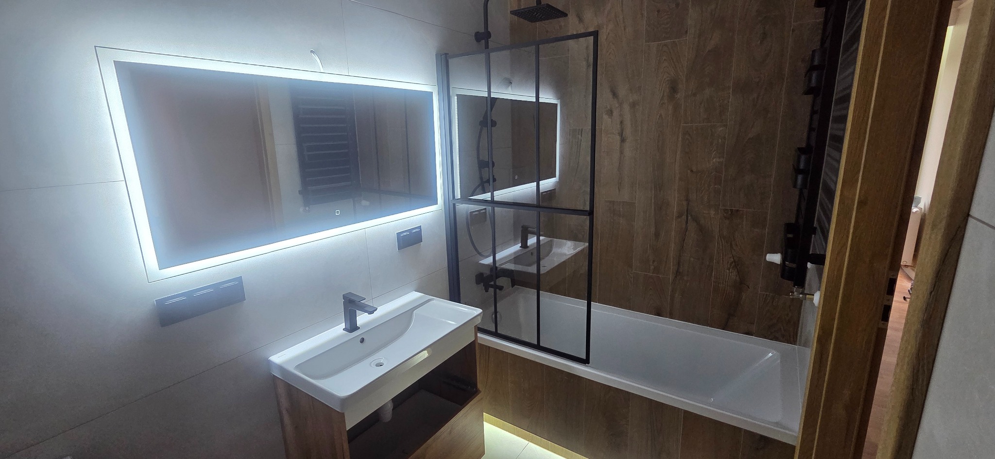 Nowoczesna łazienka z podświetlanym lustrem, kabiną prysznicową typu walk-in z czarnymi ramami i umywalką nablatową na drewnianej szafce. Płytki imitujące drewno na ścianie.