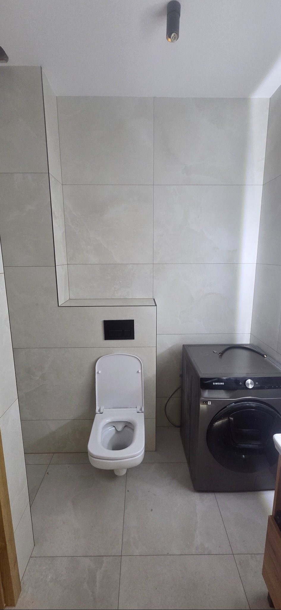 Wykończona łazienka z podwieszaną toaletą i pralką Samsung. Jasne płytki imitujące marmur na ścianach i podłodze. Minimalistyczny design, nowoczesne oświetlenie.