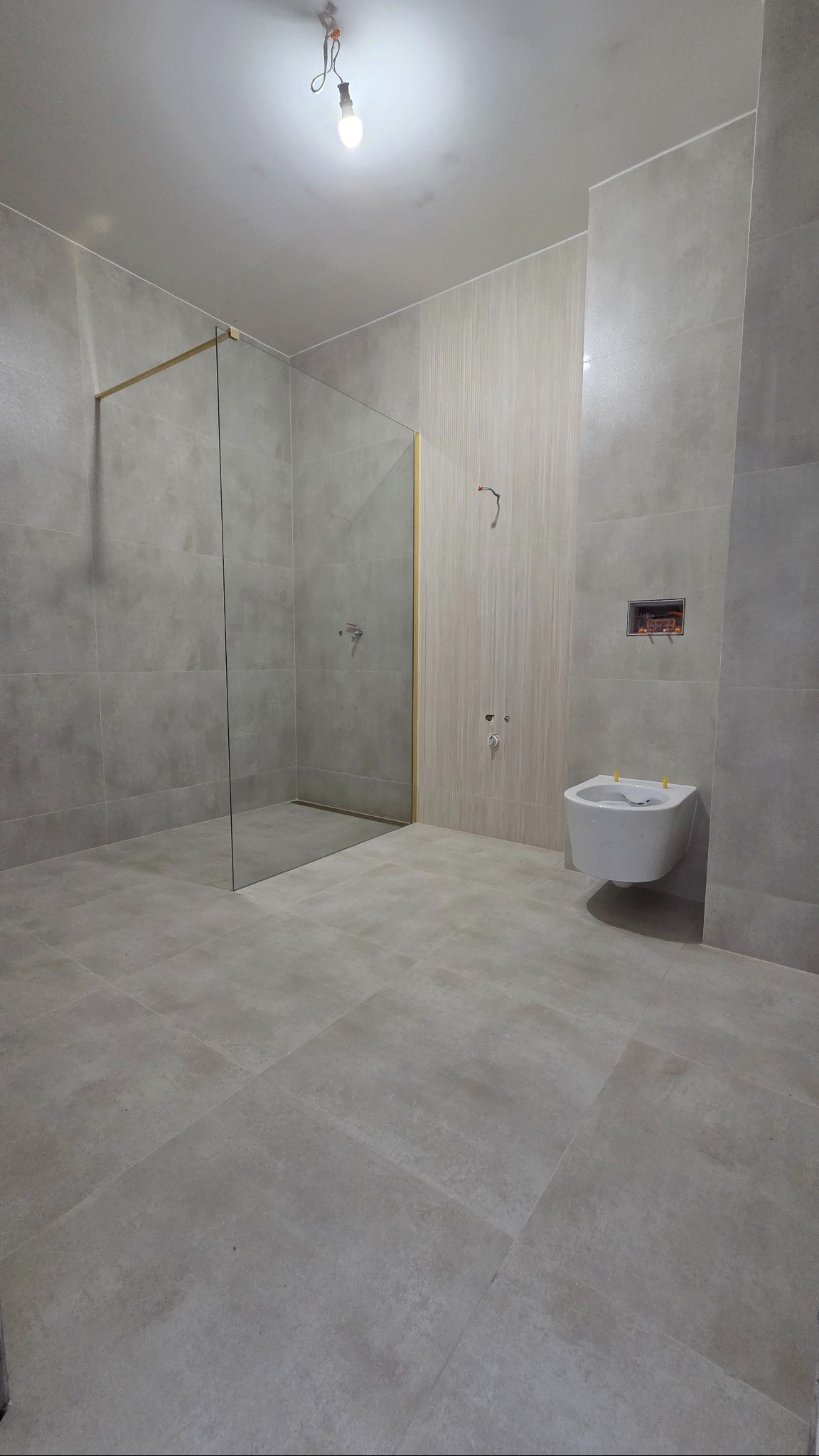 Jasna łazienka z kabiną prysznicową typu walk-in, wiszącą toaletą i beżowymi płytkami. Widoczne niedokończone elementy instalacji.