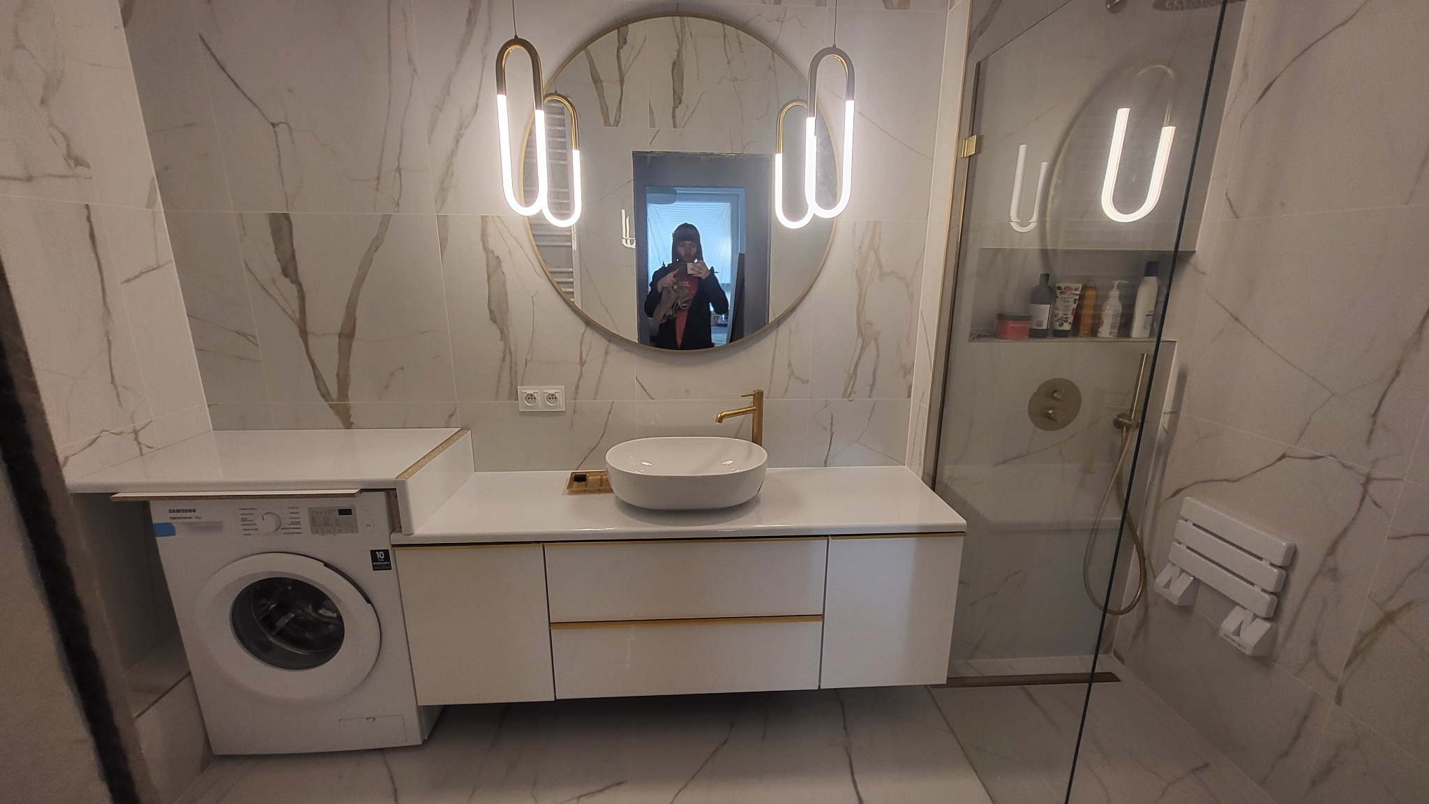 Elegancka łazienka z umywalką nablatową, złotą armaturą i okrągłym lustrem podświetlonym lampami. Kabina prysznicowa ze szklaną ścianą i siedziskiem. Płytki imitujące marmur.