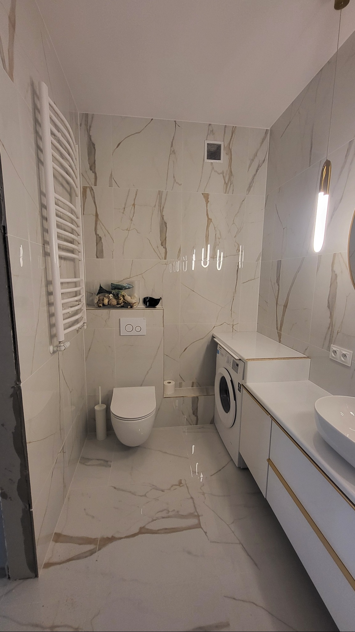 Jasna łazienka z marmurowymi płytkami, podwieszaną toaletą i grzejnikiem drabinkowym. Umywalka nablatowa i pralka w zabudowie z akcentami złota.