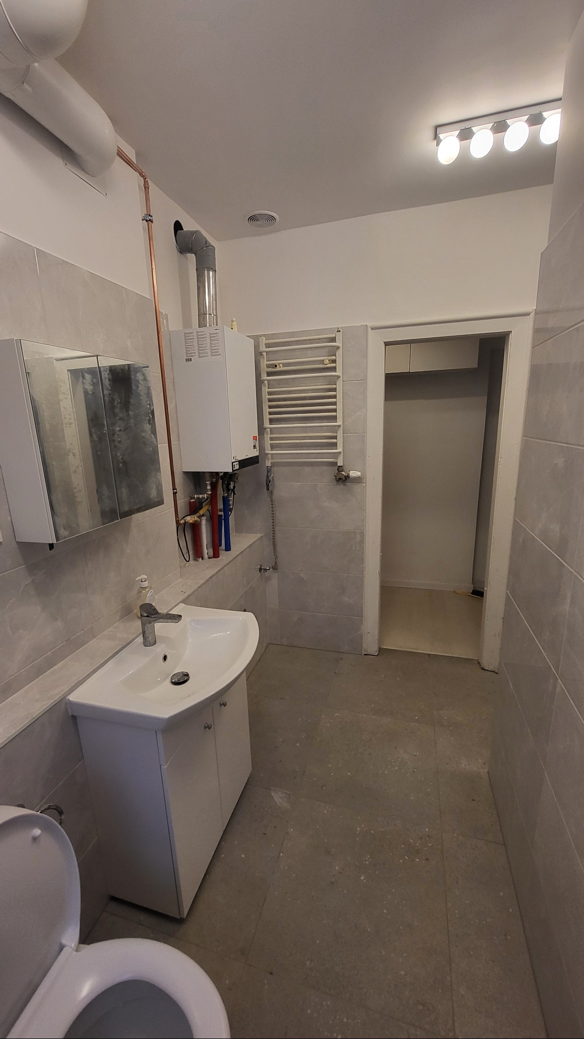 Wykończona łazienka z umywalką, szafką, grzejnikiem drabinkowym i piecem gazowym. Widoczne płytki w odcieniach szarości i biała armatura. Minimalistyczny styl.