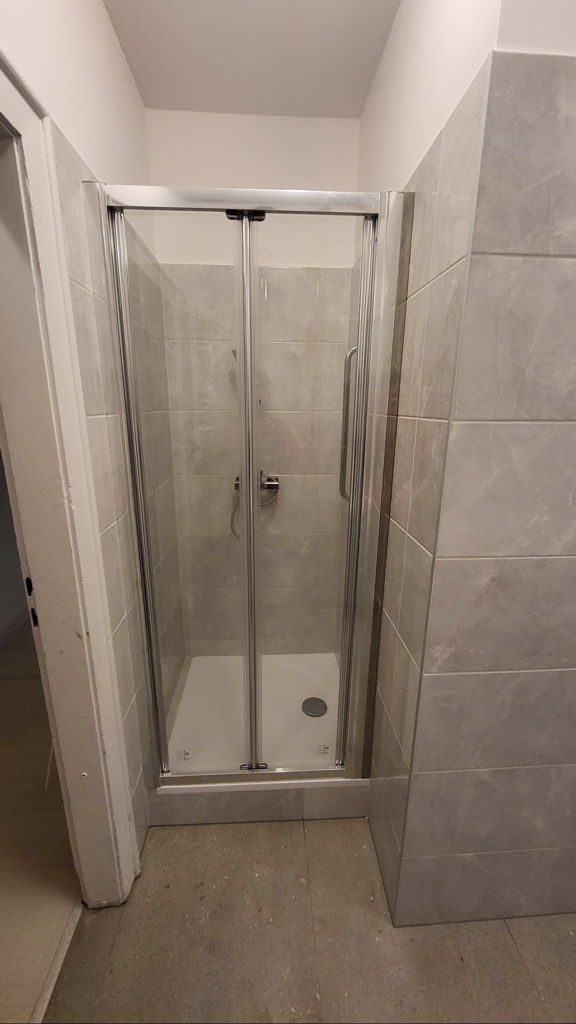 Wykończona łazienka z kabiną prysznicową ze srebrnymi ramami i szarymi płytkami imitującymi marmur na ścianach. Widoczny biały brodzik i odpływ.