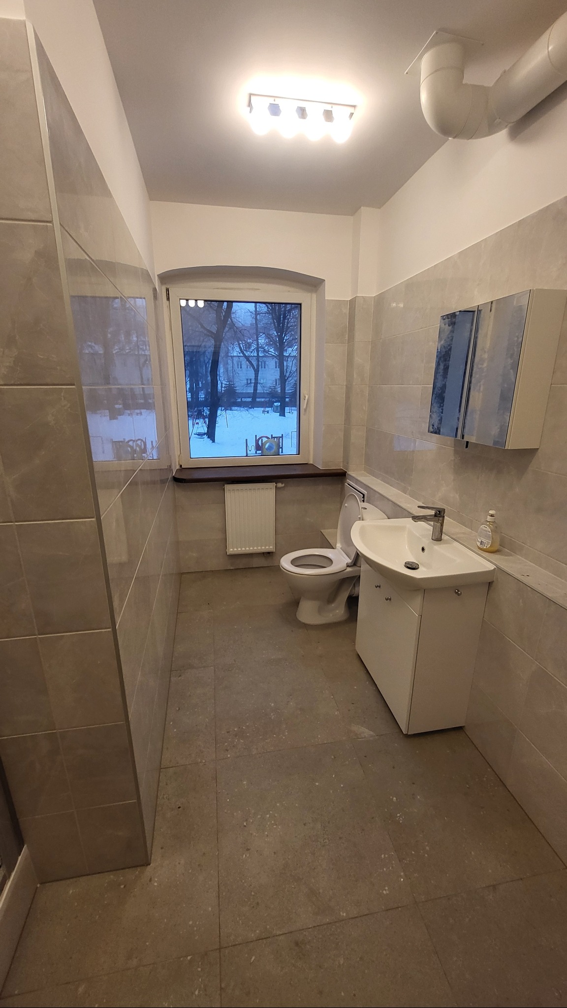 Jasna łazienka z oknem, wykończona szarymi płytkami. Umywalka z szafką, toaleta i grzejnik pod oknem. Za oknem widok na zimowy krajobraz.