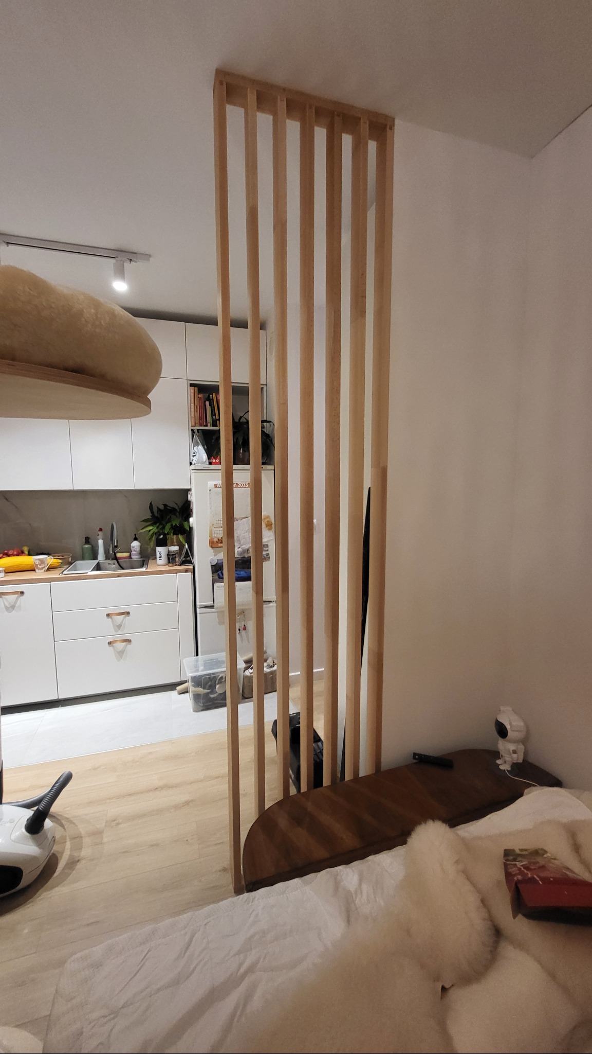 Nowoczesna, drewniana ścianka działowa z pionowych listew oddzielająca kuchnię od sypialni. Widok na białe meble kuchenne i część łóżka.