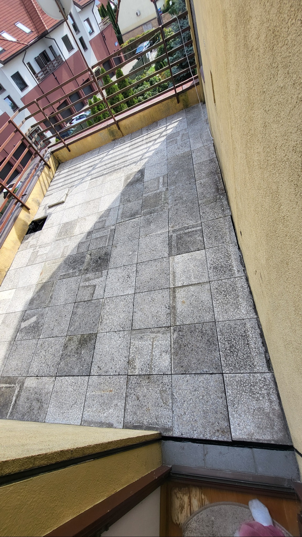 Wyłożony szarymi płytkami balkon z metalową balustradą, widok z góry. Na płytkach widoczne cienie, a w rogu balkonu otwór. Ściana balkonu w kolorze żółtym.