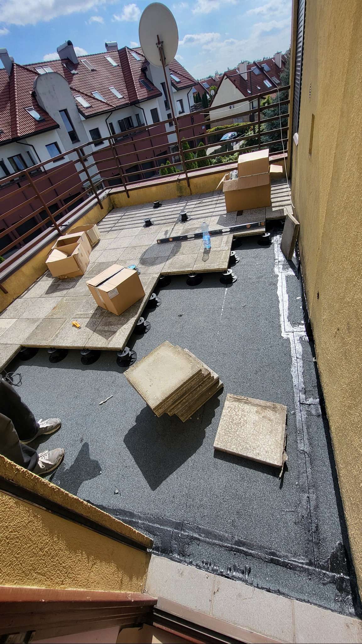 Montaż podłogi na balkonie z użyciem płyt na regulowanych wspornikach. Widoczne kartony, poziomica i częściowo ułożona podłoga. Tło stanowią budynki mieszkalne.