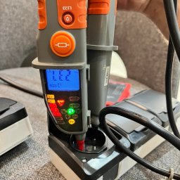 Aleks Shlosar - Pomiar napięcia w gniazdku elektrycznym za pomocą próbnika napięcia z wyświetlaczem LCD, wskazującego wartość 232V. Widoczne diody sygnalizacyjne i przewody.