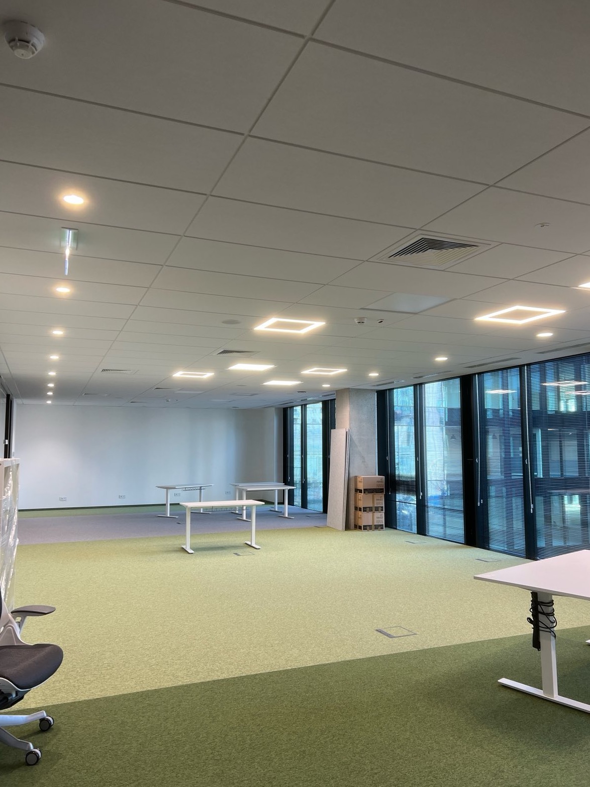 Nowoczesne biuro z kwadratowymi lampami sufitowymi LED, zieloną wykładziną i białymi biurkami. Duże okna z żaluzjami w tle. Jasne, przestronne wnętrze.