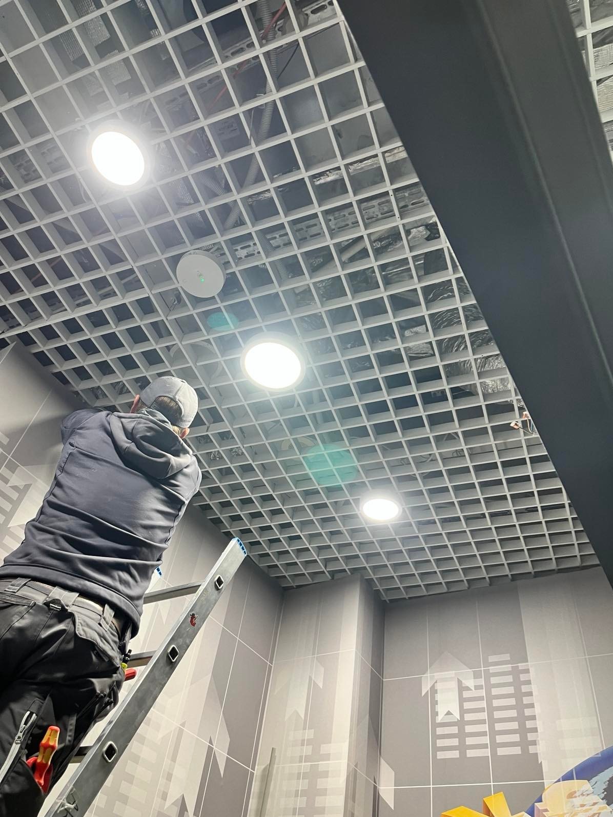 Elektryk na drabinie montuje oświetlenie w rastrowym suficie podwieszanym. Widoczne narzędzia i elementy instalacji. Nietypowy wzór na ścianie.